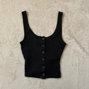 Svart ribbad linnetopp - Snygg svart ribbad linnetopp med knappar framtill. Helt oanvänd i jättebra skick, strl XSS men passar även XS (stretchig passform).