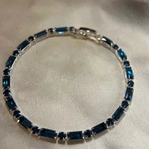 Elegant armband med blå stenar - Snyggt armband med rektangulära blå stenar infattade i silverfärgad metall. Perfekt för att ge en touch av färg och elegans till din outfit. Armbandet har en säker låsning som håller det på plats hela dagen. 17 cm lång 
