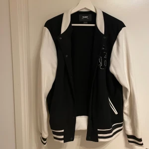Svart och vit Varsity Jacket från Pull&Bear - Snygg svart och vit Varsity Jacket från Pull&Bear med kontrasterande ärmar och randiga muddar. Jackan har knappar framtill och en stilren design som passar perfekt för en casual look.