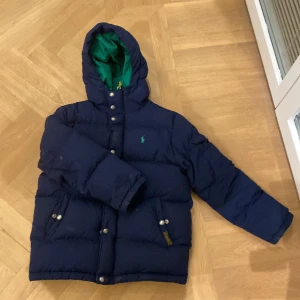 Blå dunjacka med grön insida - Säljer en snygg blå dunjacka från Polo Ralph Lauren med en grön insida. Jackan har långa ärmar och praktiska knappar framtill. Perfekt för kyliga dagar med sin pufferdesign och varma material.