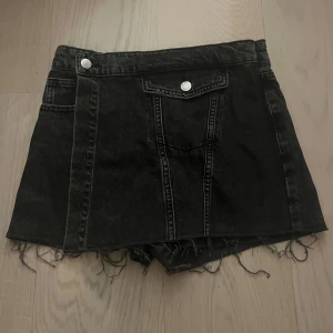 Svart jeanskjol med shorts under  med fransar - Snygg svart jeanskjol med råa fransar i nederkant och dekorativa knappar fram. Kjolen har en cool, avslappnad stil med fickor och har shorts under 