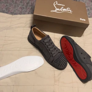 Grå sneakers från Christian Louboutin - Snygga grå sneakers från Christian Louboutin med den ikoniska röda sulan. Skorna har snörning och dekorativa nitar på tån. Perfekta för dig som vill ha en stilren och unik look. En tagg är borta