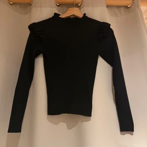 Svart ribbad tröja från ONLY - Snygg svart ribbad tröja från ONLY med volangdetaljer vid axlarna. Tröjan har långa ärmar och en hög halsringning som ger en stilren look. Perfekt för både vardag och speciella tillfällen.