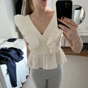 Vit blus med volanger - Säljer en söt vit blus med volanger och v-ringning. Blusen har en öppen rygg med knytning och är ärmlös. Perfekt för sommarens varma dagar.