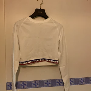 Vit croppad tröja från Tommy Hilfiger - Snygg vit croppad tröja från Tommy Hilfiger med långa ärmar. Tröjan har ett elastiskt band med Tommy Hilfiger-loggan i blått och rött vid nederkanten. Perfekt för en stilren och avslappnad look. Helt oanvänd!