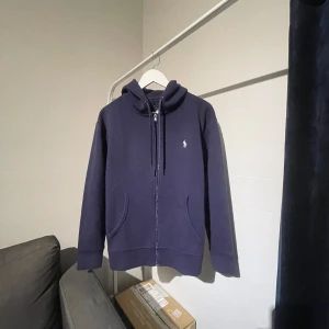 Mörkblå hoodie från Ralph Lauren - Säljer en stilren mörkblå hoodie från Ralph Lauren med dragkedja och broderad logga på bröstet. Tröjan har en klassisk passform med långa ärmar och en praktisk huva med snörning. Perfekt för en avslappnad look. Skick 9/10