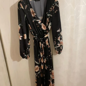 Svart blommig långklänning - Elegant svart långklänning med blommigt mönster i rosa och grönt. Klänningen har långa ärmar och en omlott-design med knytband i midjan för en smickrande passform. Storlek:xs-m. Den har små defekter här och där men inget som syns !