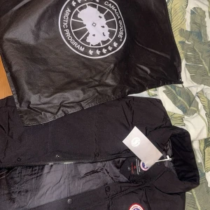 Svart canada goose väst - Säljer en svart dunjacka från Canada Goose med klassisk logga på bröstet. Jackan har en dragkedja och knappar framtill för extra skydd mot kyla. 