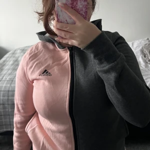 Rosa och grå huvtröja från Adidas - Säljer en snygg huvtröja från Adidas i rosa och grått. Tröjan har en dragkedja framtill och en stor logga på ryggen. Använd 1-2 gånger. 