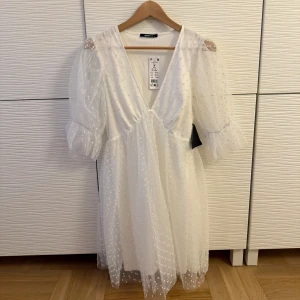 Studentklänning vit prickig  - Charmig vit klänning från Gina Tricot med prickigt mönster och v-ringad design. Klänningen har puffiga trekvartsärmar med volangdetaljer och är tillverkad i ett lätt och luftigt material. Dragkedja på sidan. Köpt för att ha som studentklänning. Storlek M. Ny med prislapp. 