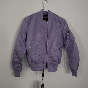 Lila bomberjacka från Alpha Industries - Snygg lila bomberjacka från Alpha Industries med dragkedja framtill och fickor med knappar. Jackan har en klassisk bomberstil med ribbade muddar och en ficka med dragkedja på ärmen. Perfekt för en cool och avslappnad look.