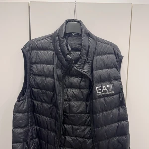 Svart dunväst från Emporio Armani - Snygg svart dunväst från Emporio Armani med EA7-logga på bröstet. Västen har en quiltad design och dragkedja.Köpt för 1100. Storlek M. Bra skick 