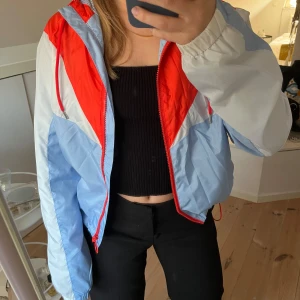 Flerfärgad vindjacka med dragkedja - Vindjacka i rött, blått och vitt med dragkedja framtill. Jackan har en luva och är justerbar i midjan. Storlek 34