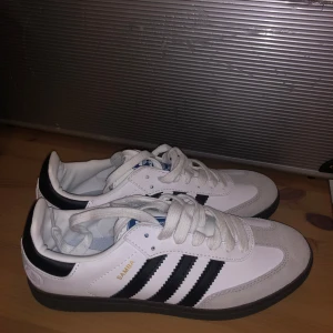 Adidas Samba sneakers - Klassiska Adidas Samba sneakers i vitt och svart med de ikoniska tre ränderna på sidan. Skorna har en svart sula och snörning framtill. Perfekta för en sportig och stilren look. Helt nya och endast testade.