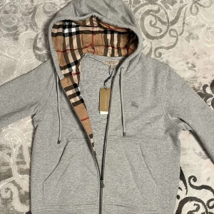 Grå hoodie från Burberry - Säljer en stilren grå hoodie från Burberry med klassiskt rutigt foder i huvan. Väldigt bra kvalitet på både zip och material. Den är helt felfri, och tags följer med! Priset är EJ hugget i sten!