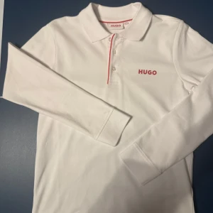 Vit långärmad pikétröja från Hugo - Snygg vit långärmad pikétröja från Hugo med röd logotyp på bröstet och röd detalj vid knäppningen. Perfekt för en stilren look.