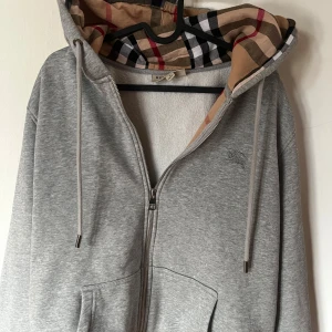 Grå hoodie från Burberry - Säljer en stilren grå hoodie från Burberry med klassiskt rutigt mönster i luvan. Tröjan har dragkedja och en broderad logga på bröstet. Perfekt för en avslappnad och trendig look. Rikigt fet Grisch !! 1  av en 1 