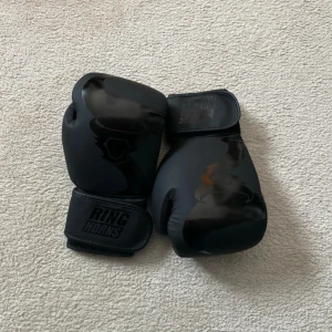 Svarta boxningshandskar från Ringhorns - Säljer ett par svarta boxningshandskar från Ringhorns. Handskarna har en stilren design med vaddering för extra skydd och komfort. Perfekta för träning och sparring. Kardborreband för enkel justering och säker passform.
