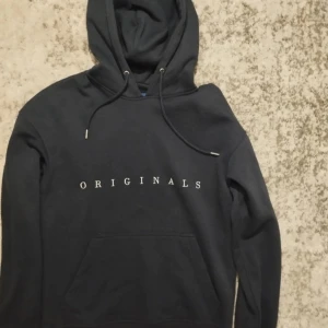 Svart hoodie med texten 'ORIGINALS' - Säljer en stilren svart hoodie med texten 'ORIGINALS' tryckt på bröstet. Den har en klassisk design med känguruficka och justerbar huva med snören. Perfekt för en avslappnad stil.