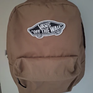 Beige ryggsäck från Vans - Säljer en beige ryggsäck från Vans med det klassiska 'Off the Wall'-märket på framsidan. Ryggsäcken har en stor ficka med dragkedja och en mindre ficka framtill. Perfekt för skolan eller vardagsbruk.
