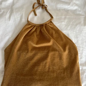 Guldglittrig halterneck topp från Ted Baker - Snygg guldglittrig halterneck topp från Ted Baker. Perfekt för en kväll ute med sitt skimrande material och eleganta design. Toppen har en knytning i nacken och en mjuk, glittrig yta som fångar ljuset fint. Mycket bra skick. Storlek 3, passar mig som vanligtvis har S.