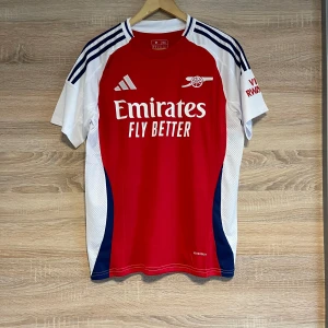 Röd Arsenal fotbollströja Saka L - Snygg röd Arsenal fotbollströja från Adidas med 'Emirates Fly Better' tryck på framsidan. Tröjan har korta ärmar och vita detaljer på sidorna samt mörkblå ränder på axlarna. På baksidan finns namnet 'Saka' och nummer 7 tryckt i vitt. Storlek L