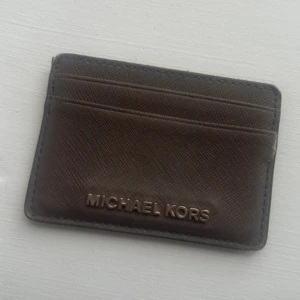 Michael Kors plånbok  - Säljer ett stilrent brunt kortfodral från Michael Kors. Perfekt för att hålla dina kort organiserade. Tillverkad i ett slitstarkt material med plats för flera kort. Se bild där det är sönder 