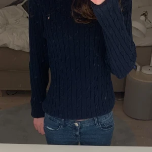 Mörkblå stickad tröja från GANT - Säljer en stilren mörkblå stickad tröja från GANT i stretchig bomull. Tröjan har ett klassiskt kabelstickat mönster och långa ärmar. Perfekt för en avslappnad men ändå elegant look.