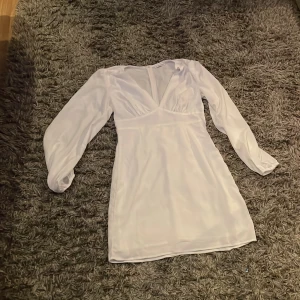 Vit klänning från Nelly x Glamorous - Elegant vit klänning med v-ringning och långa ärmar från Nelly x Glamorous. Perfekt för en stilren look med sin korta längd och figurnära passform. Klänningen har en dragkedja i sidan för enkel påklädning. Använd en gång