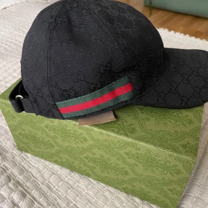 Svart keps från Gucci - Snygg svart keps från Gucci med diskret mönster och klassiska gröna och röda ränder på sidan. Justerbar rem i baktill. Str XS. Följer med tillhörande förpackning och dust påse. Förts till kvarn! Köparen står för frakten. 