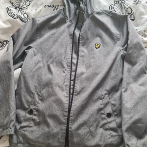 Grå vindjacka från Lyle & Scott - Snygg grå vindjacka från Lyle & Scott med dragkedja och huva. Jackan har en gul logotyp på bröstet och praktiska fickor med knappar. Perfekt för blåsiga dagar.