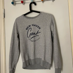 Grå sweatshirt från Gant - Säljer en stilren grå sweatshirt från Gant med tryck 'New Haven' på framsidan. Tröjan har långa ärmar och ribbade muddar vid ärmslut och nederkant. Perfekt för en avslappnad look.
