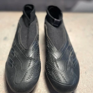 Svarta Adidas Predator fotbollsskor - Säljer ett par svarta Adidas Predator fotbollsskor i storlek 40. Skorna har en elegant design med strömlinjeformade detaljer och hög ankel för extra stöd. Yttersulan är utrustad med fasta dobbar, anpassade för spel på gräsplaner.  Skick: Begagnade men i gott skick. Yttersulan visar mindre tecken på användning, och ovandelen är välbevarad.  Nypris: 1200 SEK