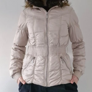 Beige jacka från H&M Divided - Säljer en trendih dunjacka från H&M Divided med pälskantad huva. Jackan har markerad midja och två fickor, stängs med dragkedja framtill. Perfekt för kyligare dagar! Pris är självklart förhandlingsbart :)
