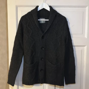 Mörkgrå stickad cardigan - Säljer en snygg mörkgrå kofta med ett diskret mönster och knappar framtill. Sjalkrage, två fickor på framsidan.