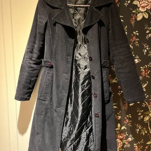 Svart manchesterkappa - Snygg svart kappa i manchester med knappar framtill och långa ärmar. Den har en klassisk krage och två fickor på sidorna. Perfekt för vår, höst och vinter. Fodrad.