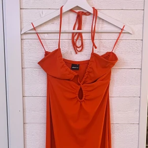 Orange klänning från Gina Tricot - Orange klänning från Gina Tricot med halterneck och knytband. Klänningen har en snygg cut-out detalj framtill och är perfekt för sommarens äventyr. Den är figurnära och har en kort längd som ger en stilren look.