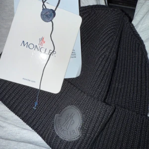 Svart ribbad mössa från Moncler - Snygg svart ribbad mössa från Moncler med ett stilrent märke framtill. Perfekt för att hålla värmen under kyliga dagar.