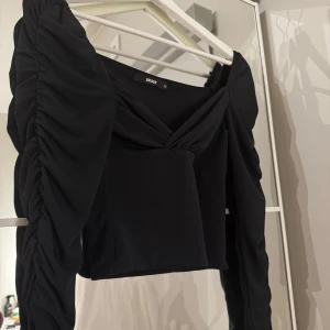 Svart topp från Bik Bok - Snygg svart topp från Bik Bok med långa ärmar och rynkade detaljer. Toppen har en omlott-design vid halsen och är croppad för en trendig look.