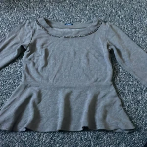 Grå armbågs längs topp med volang - Snygg grå topp med armbågs långa ärmar och volangdetalj i nederkant. Perfekt för en stilren look med en touch av elegans. Halsringningen är dekorerad med en subtil kedjedetalj.