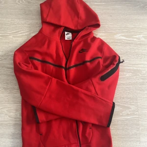 Röd huvtröja från Nike - Snygg röd huvtröja från Nike med dragkedja och svarta detaljer. Perfekt för en sportig look med sin bekväma passform och praktiska huva. Tillverkad i mjukt material för extra komfort. Den är i perfekt skick och har använts ungefär 5 ggr