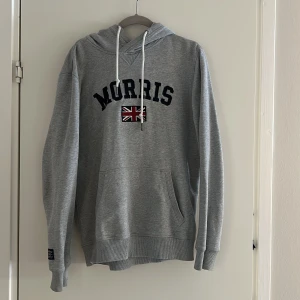 Grå hoodie från Morris - Säljer en snygg grå hoodie från Morris med tryck av Union Jack och texten 'Morris' på framsidan. Tröjan har en klassisk känguruficka och justerbar huva med snören. Perfekt för en avslappnad stil.      I storlek M.