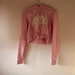 Rosa croppad hoodie från Hype - Säljer en snygg rosa croppad hoodie från Hype med text på framsidan. Perfekt för en avslappnad stil. Den har långa ärmar och en bekväm huva.