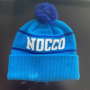 Ljusblå Nocco mössa - Snygg blå mössa från NOCCO med en stor tofs på toppen. Mössan har ribbad kant och mörkblå ränder samt vit text framtill. Perfekt för kyliga dagar.