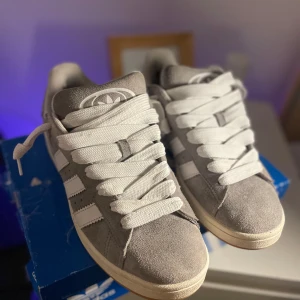  Adidas Campus - Snygga grå Adidas sneakers i mocka med vita detaljer och snörning. Perfekta för en avslappnad stil. Skorna kommer i originalkartong. Skorna är i mycket fint skick då jag går på rullstol och inte riktigt förbrukar de:) extra rep i grå färg förljer med.