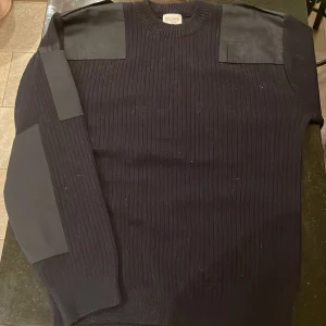 Militär ylletröja - Säljer en svart ylletröja med förstärkta axelpartier och ribbad design. Tröjan har en klassisk stil och är perfekt för kyligare dagar. Märkt med storlek 5 och passar vikt 70-80 kg. Kronomärk och är i nyskick.