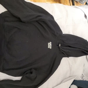 Svart hoodie från H&M med text - Säljer en svart hoodie från H&M:s Divided-kollektion med texten 'Amour Paris' broderad på bröstet. Den har en klassisk känguruficka och justerbar huva med dragsko. Perfekt för en avslappnad stil.Pris går att diskutera och köparen står för frakten