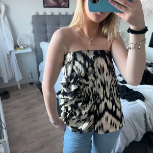 Svart och guldmönstrad tubtopp - Snygg tubtopp i svart med ett iögonfallande guldmönster. Toppen har volangdetaljer på sidan som ger en extra touch. Perfekt för en kväll ute eller en festlig tillställning. Är en kjol från början så kan användas som båda! Passar mig som är en S