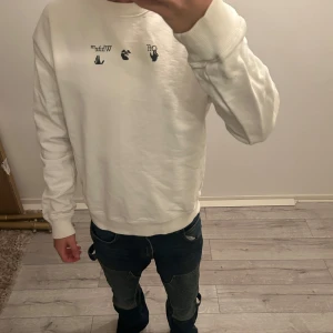 Vit sweatshirt från Off-White - Säljer en stilren vit sweatshirt från Off-White med svart text och grafik framtill. Baksidan har ett stort grönt och svart grafiskt tryck. Tröjan har långa ärmar och ribbade muddar vid ärmslut och nederkant.