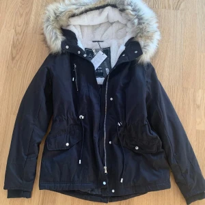 Svart parkajacka med fuskpäls storlek S - Snygg svart parkajacka med fuskpäls på huvan. Jackan har en varm fleecefodring. Helt ny.